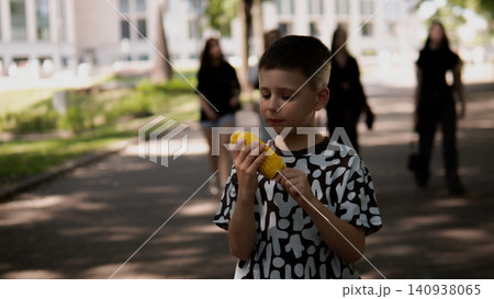 Child holding yellow corn on cob with both hands, tasting kernels while standing on tree-lined paved path in city park; soft dappled sunlight, blurred 140938065