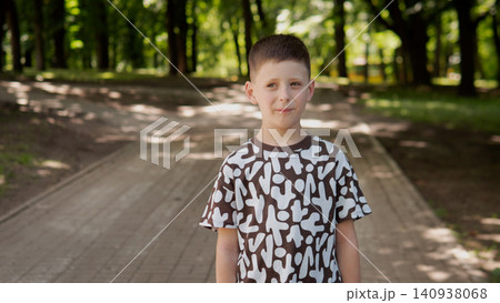 Boy standing on paved park path surrounded by mature trees and dappled sunlight, wearing short sleeve patterned t shirt and neutral shorts, calm facial Boy standing on paved park path surrounded by mature trees and dappled sunlight, wearing short sleeve patterned t shirt and neutral shorts, calm facial 140938068