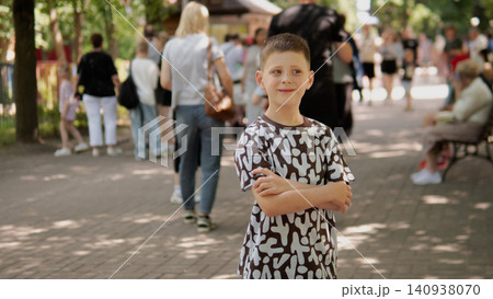 Smiling school-age boy stands with folded arms on tree-lined park walkway, wearing patterned short-sleeve shirt and shorts while people stroll and sit on Smiling school-age boy stands with folded arms on tree-lined park walkway, wearing patterned short-sleeve shirt and shorts while people stroll and sit on 140938070
