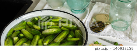 Cucumbers are soaking in a bowl while garlic and cucumber slices are on a cutting board. Several empty jars sit nearby on a clean countertop. It is a preparation activity for winter storage, banner 140938159