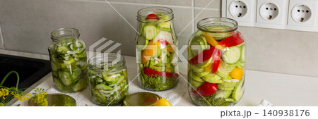 Cucumbers, peppers, and zucchini are being prepared and placed in jars for canning. Fresh ingredients are cut and organized on a clean kitchen counter for preserving, banner 140938176