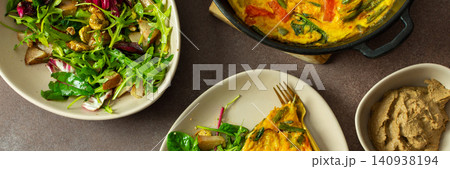 A plate features baked eggs with vegetables alongside a fresh arugula salad. Small bowls hold liver pate and baked pear pieces. This meal is set on a neutral table, banner 140938194
