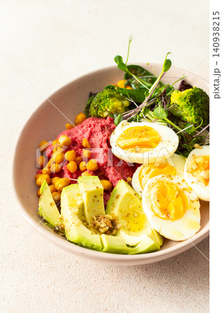 A bowl holds beet hummus topped with chickpeas, fresh broccoli, and microgreens salad alongside slices of avocado and boiled eggs. This meal is nutritious and filling 140938215