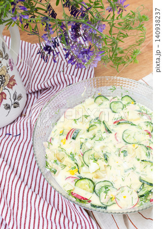 A bowl of okroshka filled with potatoes, chicken breast, cucumbers, herbs, and boiled eggs sits on a table. Nearby, a floral vase adds color to the setting A bowl of okroshka filled with potatoes, chicken breast, cucumbers, herbs, and boiled eggs sits on a table. Nearby, a floral vase adds color to the setting 140938237