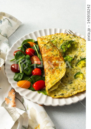 A potato omelette made with eggs and mixed greens is served on a white plate. Spinach, cherry tomatoes, and broccoli are arranged on the side for a healthy breakfast option A potato omelette made with eggs and mixed greens is served on a white plate. Spinach, cherry tomatoes, and broccoli are arranged on the side for a healthy breakfast option 140938242