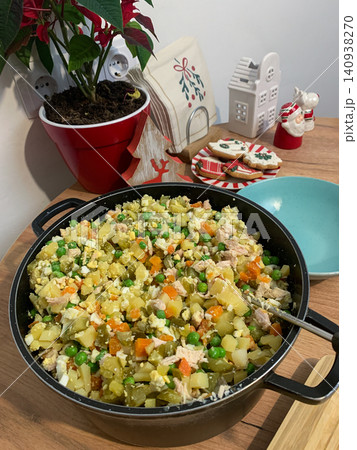 Olivier salad is being prepared in a kitchen set for Christmas and New Year. Fresh ingredients are mixed together in a large bowl on a wooden table with festive decorations 140938270