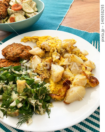 A white plate features cooked potatoes alongside fresh salad and two pancakes made from carrots and zucchini. This meal is ideal for anyone needing a nutritious breakfast or lunch A white plate features cooked potatoes alongside fresh salad and two pancakes made from carrots and zucchini. This meal is ideal for anyone needing a nutritious breakfast or lunch 140938293