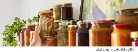 Canned vegetables and fruits are lined up on a shelf in a kitchen, showing winter preparations. A painting of a basket is seen in the background as part of the decor, banner Canned vegetables and fruits are lined up on a shelf in a kitchen, showing winter preparations. A painting of a basket is seen in the background as part of the decor, banner 140938297