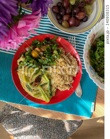 Grains, cooked greens, and vegetables are arranged on a red plate. A bowl of mixed salad with tomatoes is placed nearby on a light surface Grains, cooked greens, and vegetables are arranged on a red plate. A bowl of mixed salad with tomatoes is placed nearby on a light surface 140938315