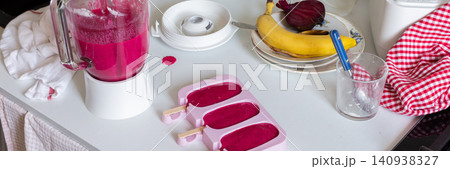 In a kitchen, a blender is mixing strawberries, bananas, and beets. Ice cream molds sit on the table filled with the mixture. A ripe banana and a beet are nearby, banner In a kitchen, a blender is mixing strawberries, bananas, and beets. Ice cream molds sit on the table filled with the mixture. A ripe banana and a beet are nearby, banner 140938327