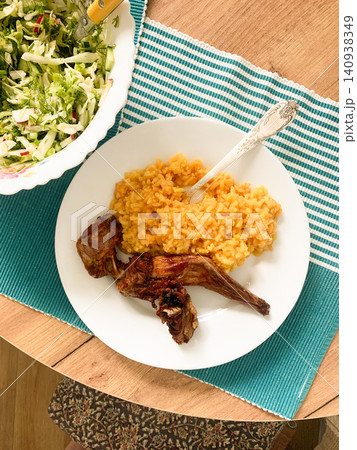 A meal is served on a white plate, showing rice colored with turmeric and pieces of baked rabbit beside a fresh salad in a bowl. The meal is set on a wooden table with a blue cloth 140938349