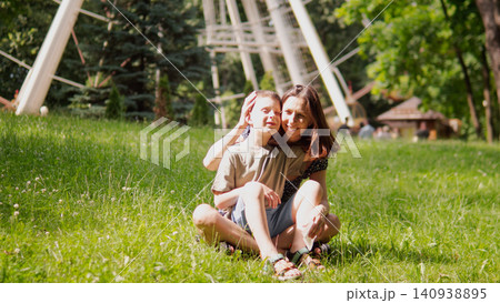 Mother and young son sitting on green grass in sunlit park, mother embracing child from behind while both smile and relax beside playground structure 140938895