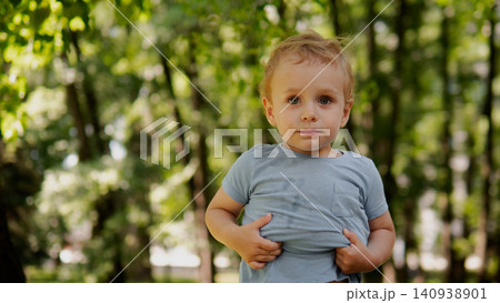 Toddler standing outdoors in leafy park, holding shirt hem with both hands and looking toward camera with curious calm expression; short light hair, blue-gray Toddler standing outdoors in leafy park, holding shirt hem with both hands and looking toward camera with curious calm expression; short light hair, blue-gray 140938901