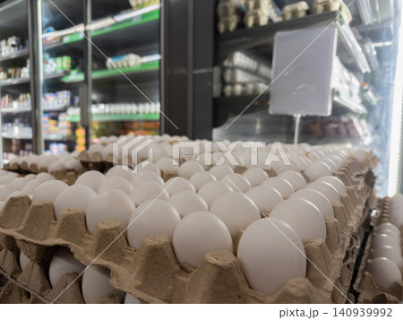 Stacked white chicken eggs in recycled cardboard crates inside a grocery store refrigeration display, emphasizing fresh organic food supply chains and daily healthy retail nutrition Stacked white chicken eggs in recycled cardboard crates inside a grocery store refrigeration display, emphasizing fresh organic food supply chains and daily healthy retail nutrition 140939992