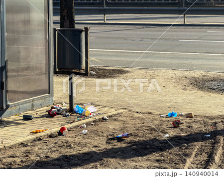 A metal waste bin stands surrounded by scattered litter on the ground at a public bus stop, depicting an urgent urban environmental problem and the need for better city maintenance A metal waste bin stands surrounded by scattered litter on the ground at a public bus stop, depicting an urgent urban environmental problem and the need for better city maintenance 140940014