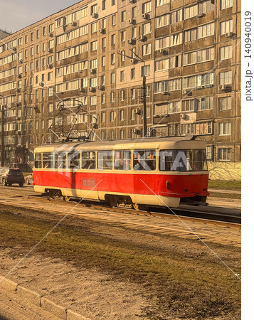 Retro red tram moving along tracks past a multi-story apartment block in a city, representing public transportation efficiency and the nostalgic charm of daily urban commuting life Retro red tram moving along tracks past a multi-story apartment block in a city, representing public transportation efficiency and the nostalgic charm of daily urban commuting life 140940019