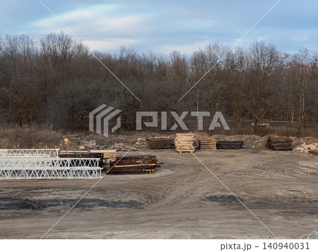 A construction site yard filled with stacks of metal beams, wooden planks, and white lattice girders set against a background of bare winter trees, symbolizing urban development A construction site yard filled with stacks of metal beams, wooden planks, and white lattice girders set against a background of bare winter trees, symbolizing urban development 140940031
