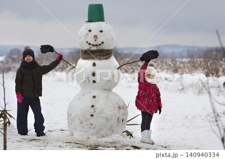 Two cute children, boy and girl, standing in front of smiling snowman in bucket hat, scarf and gloves on snowy winter landscape and blue sky copy space background. Merry Christmas, Happy New Year. Two cute children, boy and girl, standing in front of smiling snowman in bucket hat, scarf and gloves on snowy winter landscape and blue sky copy space background. Merry Christmas, Happy New Year. 140940334
