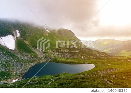 Fantastic wide peaceful view of blue lake in green valley and small tourist tents at rocky mountain with patches of snow covered with low clouds. Beauty of nature, tourism and traveling concept. 140940383