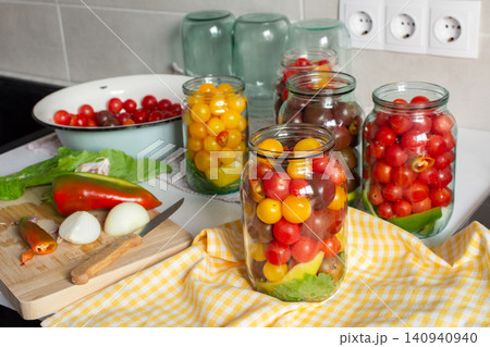 In a kitchen, jars filled with canned tomatoes sit on a table beside fresh vegetables. A mixing bowl holds whole tomatoes as preparation for winter storage continues 140940940