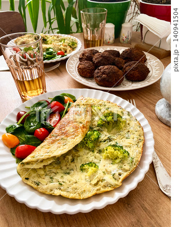 A potato omelette made with eggs and mixed greens is served on a white plate. Spinach, cherry tomatoes, and broccoli are arranged on the side for a healthy breakfast option A potato omelette made with eggs and mixed greens is served on a white plate. Spinach, cherry tomatoes, and broccoli are arranged on the side for a healthy breakfast option 140941504