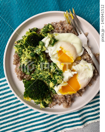 A healthy breakfast bowl features green buckwheat topped with pesto, steamed broccoli, and poached eggs. A fork rests beside the dish ready for a nutritious meal A healthy breakfast bowl features green buckwheat topped with pesto, steamed broccoli, and poached eggs. A fork rests beside the dish ready for a nutritious meal 140941512
