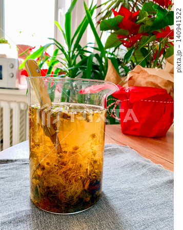 A glass container holds a decoction made from chamomile, yarrow, and corn silk. The setup includes a table, plants, and red decorations making the space inviting and bright 140941522