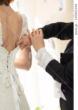 Bride in elegant white wedding dress with intricate beading adjusts her gown while a groom in a black tuxedo assists, soft lighting enhances the intimate moment Bride in elegant white wedding dress with intricate beading adjusts her gown while a groom in a black tuxedo assists, soft lighting enhances the intimate moment 140942440