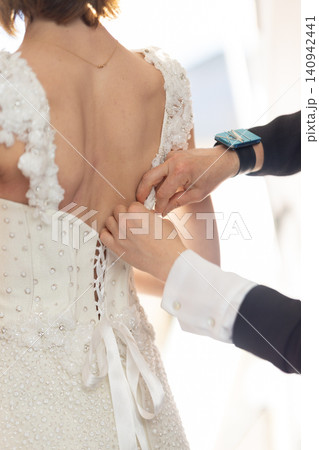 Bride in elegant white wedding dress with intricate beadwork having her gown fastened by a helper, soft lighting highlighting the delicate details of the attire Bride in elegant white wedding dress with intricate beadwork having her gown fastened by a helper, soft lighting highlighting the delicate details of the attire 140942441