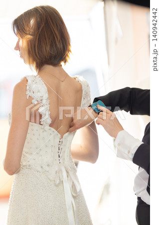 Bride in elegant white wedding dress with lace details being assisted by a person in formal attire while preparing for the ceremony in a bright indoor setting Bride in elegant white wedding dress with lace details being assisted by a person in formal attire while preparing for the ceremony in a bright indoor setting 140942442