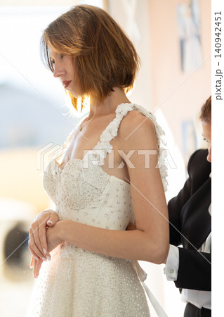 Bride in elegant white wedding dress with floral embellishments being assisted by a stylist in a bright dressing room with soft natural light Bride in elegant white wedding dress with floral embellishments being assisted by a stylist in a bright dressing room with soft natural light 140942451