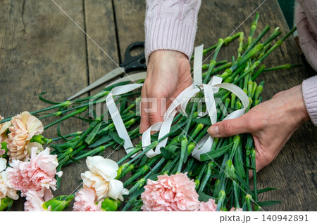 A person arranges pink flowers on a wooden table while tying them with ribbons in a bright workspace A person arranges pink flowers on a wooden table while tying them with ribbons in a bright workspace 140942891