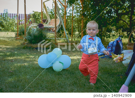 smiling boy 1 year old boy dressed in a blue shirt red bowtie and pants with a wooden camera on his neck in a green garden is barefoot on the grass, a boy of European appearance with blue eyes. smiling boy 1 year old boy dressed in a blue shirt red bowtie and pants with a wooden camera on his neck in a green garden is barefoot on the grass, a boy of European appearance with blue eyes. 140942909