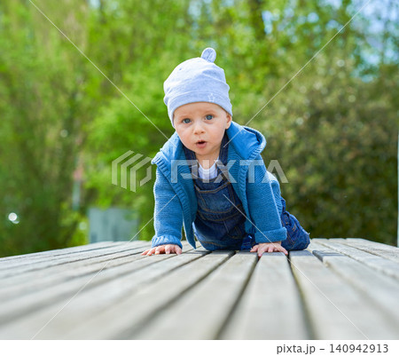 boy up to one year old boy European appearance blue eyes sits in summer on a wooden bench dressed in denim overalls and a light cap . High quality photo 140942913