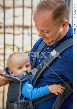 a boy under one year old sits at his father's side in a kangaroo backpack and looks into the camera, European-looking blondes with blue eyes, a baby with a pacifier in his mouth. High quality photo a boy under one year old sits at his father's side in a kangaroo backpack and looks into the camera, European-looking blondes with blue eyes, a baby with a pacifier in his mouth. High quality photo 140942933