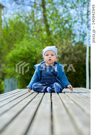 boy up to one year old boy European appearance blue eyes sits in summer on a wooden bench dressed in denim overalls and a light cap . High quality photo 140942949
