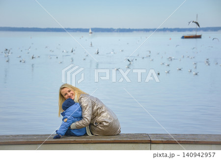 A mother holds her one-year-old son on her lap, they sit with their backs to the camera and look out over the lake, over which seagulls fly and a small sailboat sails on a fall day. High quality photo A mother holds her one-year-old son on her lap, they sit with their backs to the camera and look out over the lake, over which seagulls fly and a small sailboat sails on a fall day. High quality photo 140942957