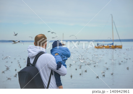 A father holds his one-year-old son in his arms, they stand with their backs to the camera and look at the lake over which seagulls fly and a small sailboat sails on an autumn day. High quality photo 140942961