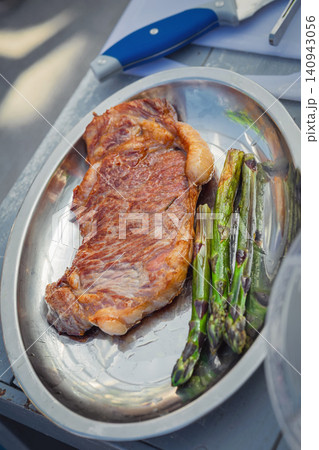 Grilled steak with asparagus is served on metal plate during outdoor cooking. Top view. Close-up of meat and vegetables prepared at food event Grilled steak with asparagus is served on metal plate during outdoor cooking. Top view. Close-up of meat and vegetables prepared at food event 140943056