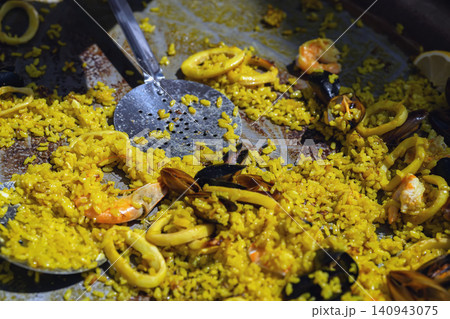 Paella fills pan with saffron rice, squid, shrimp, mussels, and serving spoon. Paella represents Spanish cuisine, seafood meal, catering, market food, restaurant menu, Mediterranean cooking Paella fills pan with saffron rice, squid, shrimp, mussels, and serving spoon. Paella represents Spanish cuisine, seafood meal, catering, market food, restaurant menu, Mediterranean cooking 140943075