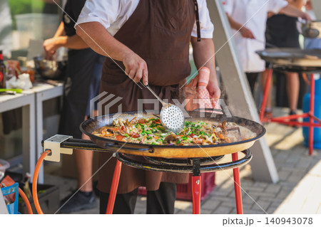 Chef cooks paella in pan with seafood, rice, herbs, steam during outdoor catering. Paella connects Spanish cuisine, street food, festival dining, restaurant service, gastronomy Chef cooks paella in pan with seafood, rice, herbs, steam during outdoor catering. Paella connects Spanish cuisine, street food, festival dining, restaurant service, gastronomy 140943078