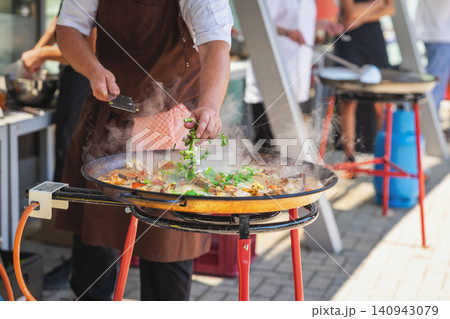 Chef cooking traditional Spanish paella in a large pan with seafood and fresh herbs at an outdoor food festival. Steam rises from hot rice dish during live cooking Chef cooking traditional Spanish paella in a large pan with seafood and fresh herbs at an outdoor food festival. Steam rises from hot rice dish during live cooking 140943079