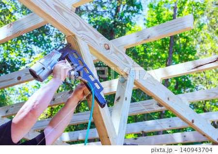 Worker connects wooden beams using power air gun in construction project surrounded by trees Worker connects wooden beams using power air gun in construction project surrounded by trees 140943704