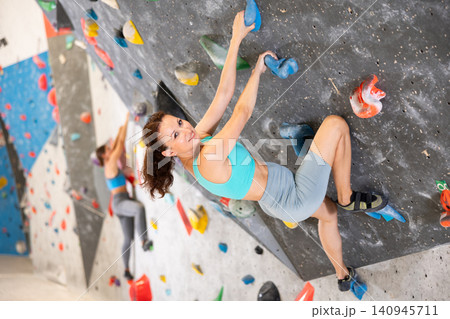 Woman in comfortable athletic clothing moving carefully on bouldering wall clinging to colorful hand and foot holds 140945711