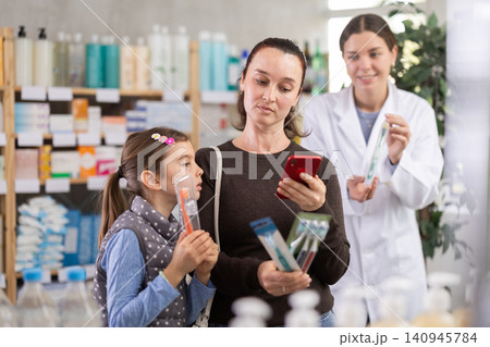 Woman with daughter in pharmacy scans QR code on toothbrush kit. Pharmacist in background Woman with daughter in pharmacy scans QR code on toothbrush kit. Pharmacist in background 140945784