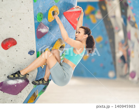 Woman in comfortable athletic clothing moving carefully on bouldering wall clinging to colorful hand and foot holds 140945983