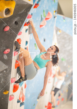 Adult woman is mastering climbing on training wall in gym, side view. Young woman holds on tightly to ledges and strives for top of bouldering route Adult woman is mastering climbing on training wall in gym, side view. Young woman holds on tightly to ledges and strives for top of bouldering route 140946005
