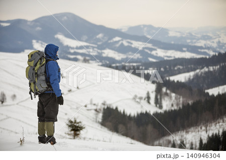 Back view of tourist hiker in warm clothing with backpack standing on mountain clearing on copy space background of woody mountain ridge and cloudy sky. Back view of tourist hiker in warm clothing with backpack standing on mountain clearing on copy space background of woody mountain ridge and cloudy sky. 140946304