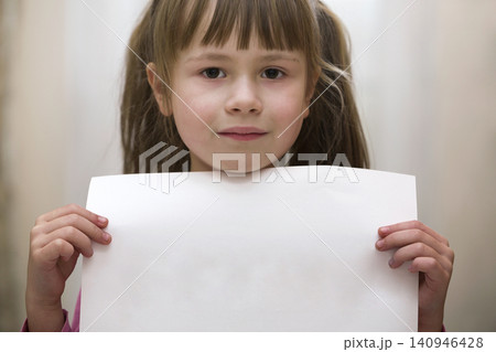 Happy smiling cute child girl holding white copy space sheet of drawing paper. Art education, creativity, advertisement concept. Happy smiling cute child girl holding white copy space sheet of drawing paper. Art education, creativity, advertisement concept. 140946428