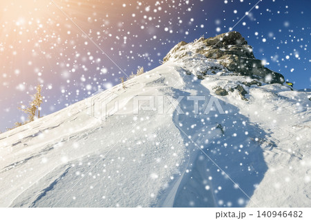 Beautiful winter landscape. Steep mountain hill slope with white deep snow, distant mountain range panorama, large snowflakes and bright shining sun rays on blue sky colorful copy space background. Beautiful winter landscape. Steep mountain hill slope with white deep snow, distant mountain range panorama, large snowflakes and bright shining sun rays on blue sky colorful copy space background. 140946482